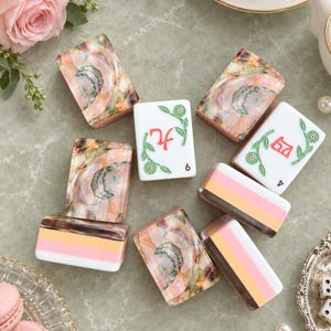 Könnte beinhalten: Eine Sammlung rechteckiger Mahjong-Spielsteine mit einer Mischung aus irisierenden Abalone-Muschelmustern, weißen Oberflächen mit grünen Blumenmotiven und roten chinesischen Schriftzeichen sowie geschichteten Blöcken aus orangefarbenem, rosa und weißem Acryl.