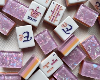 May include: A collection of Mahjong tiles featuring a mix of white tiles with engraved bamboo, flower, and joker symbols in purple and gold, alongside translucent purple tiles with iridescent flecks and layered orange and white bases.