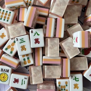 May include: A collection of mahjong tiles featuring a mix of designs, including pineapple patterns, Chinese characters, and numbers. Some tiles have a solid white face with colorful graphics, while others display a translucent, pearlescent finish.