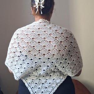 Peut inclure: Un châle triangulaire de couleur crème présentant un motif complexe de points de crochet en éventail avec des détails ajourés. Le vêtement est drapé sur les épaules, mettant en valeur une texture délicate en dentelle et un bord inférieur pointu.