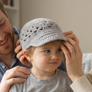 Puede incluir: Una gorra de ganchillo de color gris claro con una visera corta y un patrón calado decorativo alrededor de la corona. El gorro está confeccionado con hilo y presenta un diseño de punto texturizado hecho a mano que cubre la cabeza.