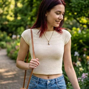 Puede incluir: Un top corto de ganchillo color crema de manga corta con cuello redondo, combinado con vaqueros azules y un bolso bandolera redondo de paja trenzada con una fina correa de cuero marrón, usado con un sencillo collar con colgante plateado.