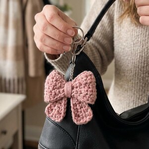 Puede incluir: Un llavero de lazo de ganchillo en color rosa empolvado sujeto a un bolso negro. El lazo está hecho de hilo grueso con un patrón de puntos texturizados y anudados, con un detalle central envuelto, unido a una anilla y cierre de metal plateado.