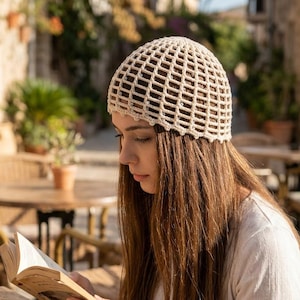May include: A cream-colored crochet beanie featuring a grid-like openwork pattern. The hat is constructed from yarn with a scalloped edge along the bottom hem, providing a textured, lightweight appearance that fits snugly around the crown of the head.