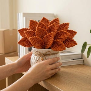 May include: A white ceramic pot with a textured vertical ribbed pattern, tied with a thin jute twine bow, containing a cluster of numerous burnt orange crocheted leaves arranged to resemble a plant with a dense, rounded foliage structure.