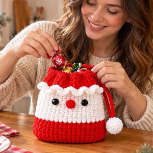 Pode incluir: Uma bolsa de crochê vermelha e branca com cordão, desenhada para se parecer com um rosto de Papai Noel, com acabamento em babados brancos, nariz vermelho, dois olhos pretos e uma ponta de gorro vermelha pendente com um pompom branco.