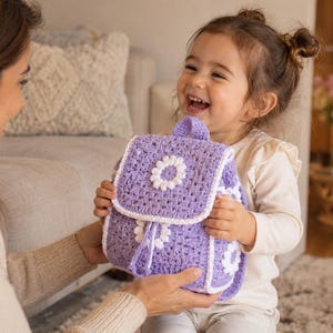 May include: A lavender crochet backpack featuring a white daisy motif on the front flap and body. The bag is constructed with a square-patterned knit texture, a top handle, and a drawstring closure, finished with a white border along the edges.