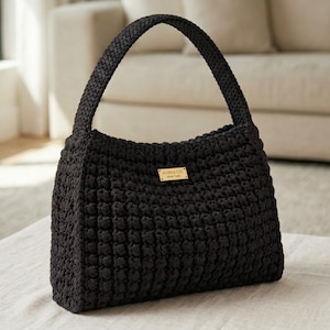 May include: A black crochet handbag featuring a textured bobble stitch pattern and a single integrated handle. A small rectangular gold-toned metal plaque is centered on the front, engraved with the text MARIA LIZ NEW YORK in black lettering.