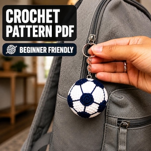 May include: A crochet soccer ball keychain attached to a grey backpack zipper. The ball features a classic white and dark blue pentagonal pattern. Text on the image reads CROCHET PATTERN PDF and BEGINNER FRIENDLY with a small yarn ball icon.