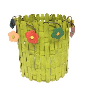 May include: A green woven planter with a decorative fence design and colorful flower accents. The planter is made of metal and has a distressed finish.