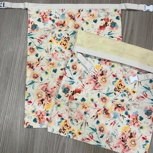 Pode incluir: Duas bolsas retangulares de tecido com fundo creme e um padrão floral colorido em tons de rosa, amarelo, laranja e azul-petróleo. Cada bolsa tem uma borda superior bege com uma alça ajustável e uma fivela de plástico branca.