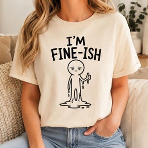 May include: A cream-coloured short-sleeve t-shirt featuring the black text "I'M FINE-ISH" above a graphic of a melting, tired-looking stick figure character giving a thumbs-down gesture, with drips falling from its body onto the ground.