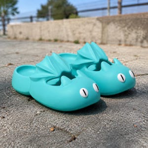 Peut inclure: Une paire de sandales à enfiler bleu turquoise vif avec un motif de dragon moulé. La chaussure présente des ailes sculptées sur les côtés, deux petites cornes à l'avant et des yeux ovales blancs avec des pupilles verticales noires.