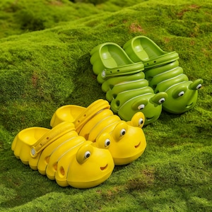 May include: A pair of bright green and a pair of bright yellow slip-on clogs shaped like caterpillars. Each shoe features a segmented body design, a rounded toe with two protruding eyes, and a small smiling mouth detail on the front section.