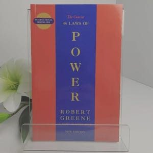 May include: A paperback book titled The Concise 48 Laws of Power by Robert Greene, featuring a vertical blue stripe on a coral background. The cover includes the text International Bestseller and New Edition, displayed in a clear acrylic stand.