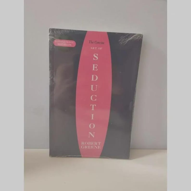 May include: A black book titled The Concise Art of Seduction by Robert Greene, featuring a prominent pink vertical oval shape in the centre with white text. The cover includes a small pink rectangular label stating International Bestseller.