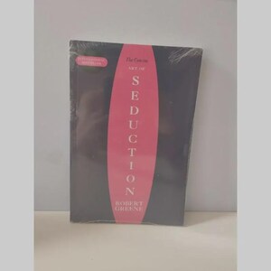 May include: A black book titled The Concise Art of Seduction by Robert Greene, featuring a prominent pink vertical oval shape in the centre with white text. The cover includes a small pink rectangular label stating International Bestseller.