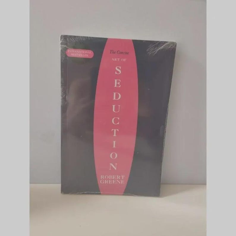 May include: A black book titled The Concise Art of Seduction by Robert Greene, featuring a prominent pink vertical oval shape in the centre with white text. The cover includes a small pink rectangular label stating International Bestseller.