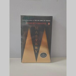 May include: A paperback book titled Mastery by Robert Greene, featuring a black cover with two large gold triangles forming an M shape. The text reads Bestselling author of THE 48 LAWS OF POWER, Robert Greene, Mastery, and The #1 New York Times Bestseller.