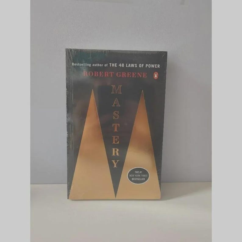May include: A paperback book titled Mastery by Robert Greene, featuring a black cover with two large gold triangles forming an M shape. The text reads Bestselling author of THE 48 LAWS OF POWER, Robert Greene, Mastery, and The #1 New York Times Bestseller.