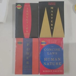 May include: Four books by Robert Greene arranged in a grid. Top left: Art of Seduction with a red oval on black. Top right: Mastery with gold triangles on black. Bottom left: The 48 Laws of Power in red and blue. Bottom right: The Concise Laws of Human Nature.