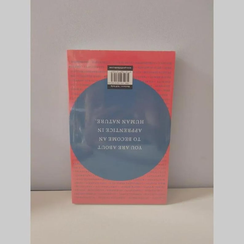 May include: A rectangular book with a coral-pink cover featuring a large central blue circle. The text reads: YOU ARE ABOUT TO BECOME AN APPRENTICE IN HUMAN NATURE. A barcode and website URL are printed in a small black box at the top of the cover.
