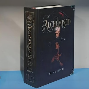 Puede incluir: Un libro de tapa dura titulado Alchemised de Senlinyu con una cubierta negra que presenta una ilustración de dos figuras. El lomo muestra el título en letras doradas con bordes decorativos y protectores de esquina de metal plateado.