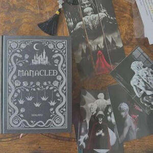 May include: A black hardback book titled Manacled by Senlinyu with white line art of a castle, snakes, roses, and origami cranes on the cover, accompanied by several black and white art prints featuring figures and a red cloak.