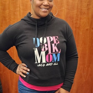 May include: A woman wearing a black hooded sweatshirt with the text "Dope Blk Mom" and "Jack and Jill" in pink, blue, and white lettering. She is wearing blue jeans and has short brown hair.