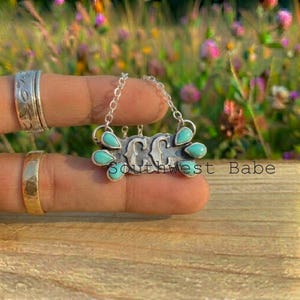 Puede incluir: Un collar con colgante de plata que presenta las letras CC centradas entre seis piedras de turquesa en forma de lágrima. El colgante cuelga de una delicada cadena de plata. El diseño incluye un acabado metálico texturizado alrededor de las piedras.