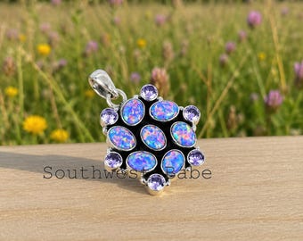 May include: A silver pendant featuring five oval-shaped blue opals and six round purple gemstones arranged in a cluster. The metal setting is polished silver. The text "Southwest Babe" is printed in black letters across the bottom of the pendant.