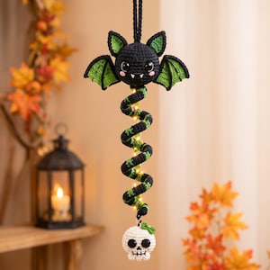May include: A hanging crochet decoration featuring a black bat with green-lined wings, large black eyes, and white fangs. Below the bat is a black and green spiral tail wrapped in thin string lights, ending with a small white crochet skull.