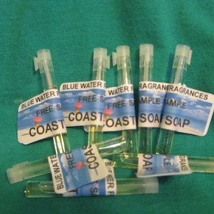 May include: Six clear glass vials with yellow liquid inside. Each vial has a label with the text "Free Sample" and the name of a fragrance. The labels are blue, white, and yellow with a beach scene and an ice cream cone.