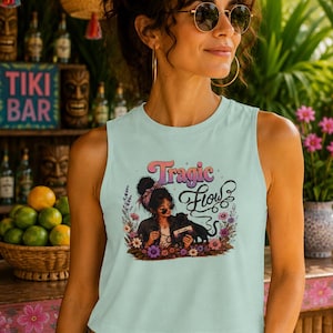 May include: A light mint green sleeveless crop top featuring a graphic print of a person with a black cat, surrounded by colorful flowers. The text "Tragic Flow" is printed above the illustration in a stylized, retro-inspired pink and purple font.