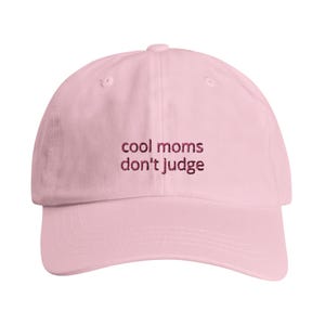 Könnte beinhalten: Eine hellrosa Baseballkappe mit dem gestickten Text "cool moms don't judge" in dunkelrosa Garn, der mittig auf der Vorderseite platziert ist. Die Kappe hat eine strukturierte Krone, einen gebogenen Schirm und einen Knopf oben.