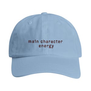Könnte beinhalten: Eine hellblaue Baseballkappe mit dem gestickten Text "main character energy" in dunkelroter Schrift, der mittig auf der Vorderseite platziert ist. Die Kappe hat eine strukturierte Krone, einen gebogenen Schirm und einen Knopf oben.