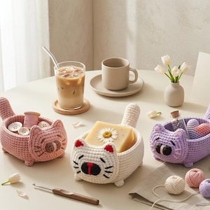 May include: Three small, hand-crocheted cat-shaped baskets in pink, cream, and lavender. Each basket features a cat face with embroidered details, a tail, and four small feet. The baskets hold various items like buttons, soap, and small hearts.
