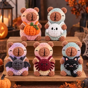 May include: Five crocheted bear plushies in various pastel colours, each wearing a different Halloween-themed accessory: an orange pumpkin, a white ghost, a grey bat, a dark red spider, and a black cat. The bears have brown muzzles and ears.
