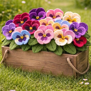 May include: A rectangular wooden planter box with rope handles containing a collection of crocheted pansy flowers in shades of purple, pink, blue, yellow, and red, accompanied by textured green crocheted leaves arranged in a dense cluster.