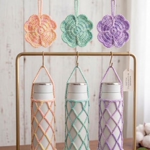 May include: Three white metal water bottles encased in pastel peach, mint green, and lavender crocheted net holders with matching flower-shaped crochet ornaments hanging above on a gold-toned metal display stand with a wooden base.