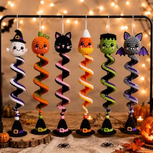 Może przedstawiać: Sześć wiszących szydełkowych ozdób na Halloween przedstawiających ducha, dynię, czarnego kota, cukierek kukurydziany, potwora Frankensteina i nietoperza. Każda figurka znajduje się na kolorowym spiralnym ogonie zakończonym podstawą w kształcie kapelusza.