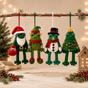 May include: Four crocheted frog ornaments hanging from a branch. The first is a Santa frog with a red hat and white beard. The second is an elf frog in green and red. The third is a snowman frog with a black hat. The fourth is a tree frog.