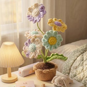 May include: A crocheted flower arrangement in a brown pot featuring five multi-colored flowers with pastel petals in shades of purple, yellow, and light blue, supported by green stems and leaves, displayed alongside a small lamp and notebook.