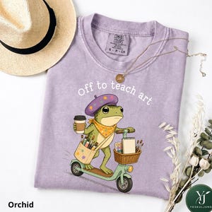 May include: A light purple t-shirt featuring a graphic of a green frog wearing a purple beret, riding a scooter, and holding a coffee cup. The frog carries a bag of paintbrushes and a basket with a sketchbook. The text reads, off to teach art.