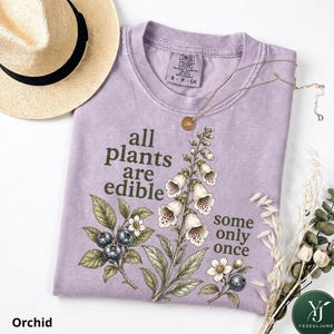 May include: A lavender t-shirt featuring a botanical illustration of blueberries and foxglove flowers with the text "all plants are edible some only once". The shirt has a crew neckline and a Comfort Colors label. A straw hat is positioned nearby.