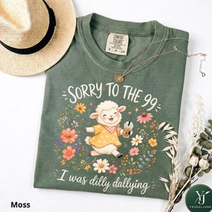 May include: A moss green t-shirt featuring a graphic of a white lamb wearing a yellow polka dot shirt and holding an iced coffee, surrounded by colorful flowers. The text reads SORRY TO THE 99 I was dilly dallying. A straw hat and necklace are present.