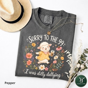 May include: A charcoal gray t-shirt featuring a graphic of a white lamb wearing a yellow polka dot shirt and holding a drink. The text reads "SORRY TO THE 99" and "I was dilly dallying" surrounded by colorful flowers. A gold necklace rests on the shirt.