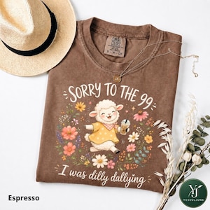 May include: A brown t-shirt featuring a graphic of a white lamb wearing a yellow polka dot dress and holding an iced coffee, surrounded by colorful flowers. The text reads SORRY TO THE 99 and I was dilly dallying. A gold necklace rests on the collar.