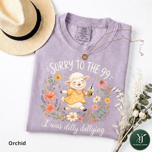 May include: A light purple t-shirt featuring a graphic of a lamb wearing a yellow polka dot dress and holding a drink, surrounded by colorful flowers. The text reads "SORRY TO THE 99" and "I was dilly dallying". A straw hat and gold necklace are included.