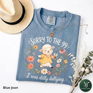 May include: A blue t-shirt featuring a graphic of a white lamb wearing a yellow polka dot dress and holding a coffee cup, surrounded by colorful flowers. The text reads SORRY TO THE 99 and I was dilly dallying. A straw hat and necklace are included.